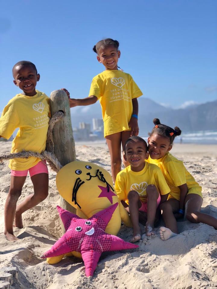 Four children in yellow Darcey Sunshine Foundation t-shirts on a beach with plush toys.