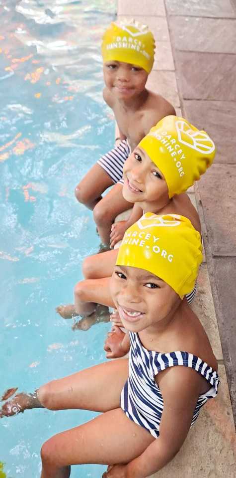 Three children wearing yellow Darcey Sunshine Foundation swim caps sit at the edge of a pool.