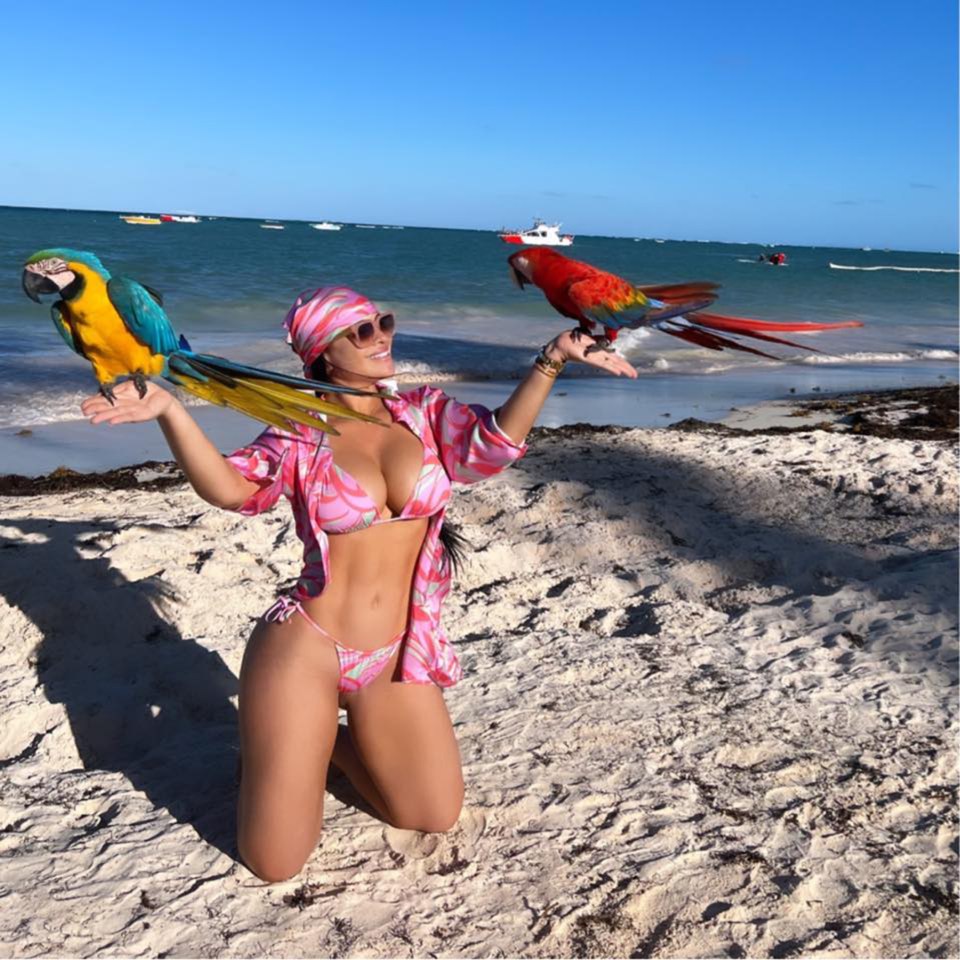 Woman in bikini holding two macaws on a beach.