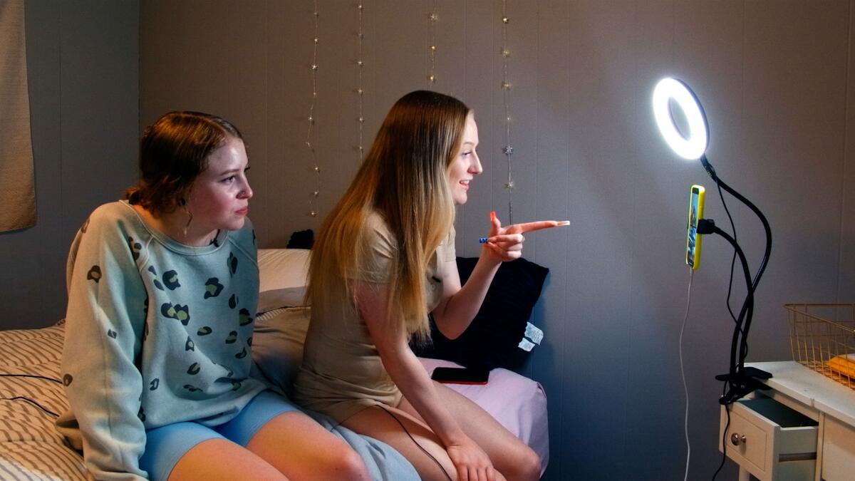 Two women illuminated by a ring light sit on a bed talking into a cellphone