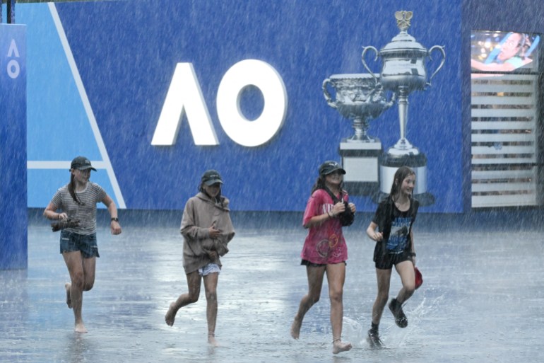 Youths run barefoot in heavy rain beside tournament signage on day one of the Australian Open tennis tournament in Melbourne on January 12, 2025. (Photo by WILLIAM WEST / AFP) / -- IMAGE RESTRICTED TO EDITORIAL USE - STRICTLY NO COMMERCIAL USE --