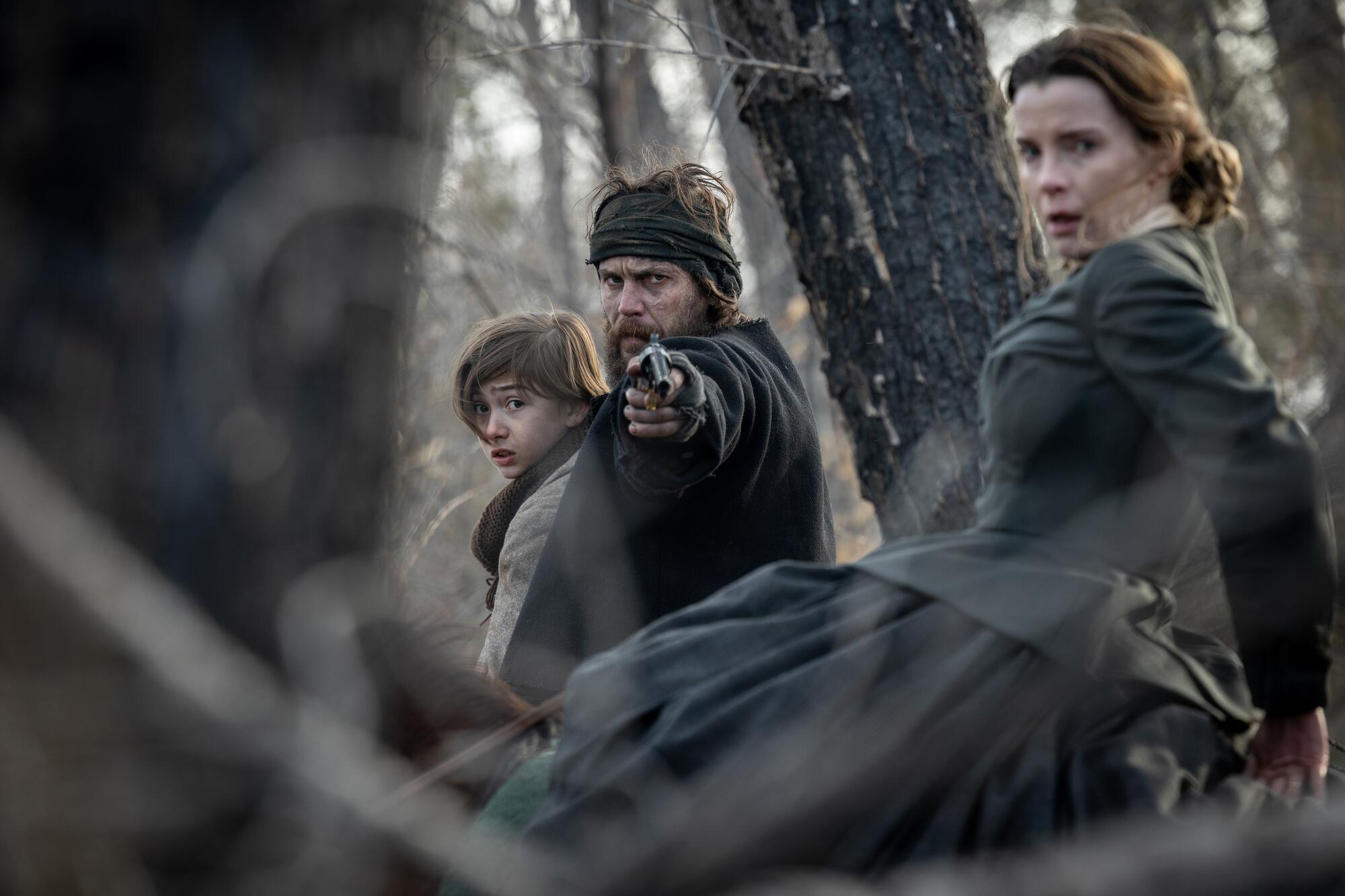 A frightened boy held by a man pointing a gun in front of him as a woman in black dress sits near them.