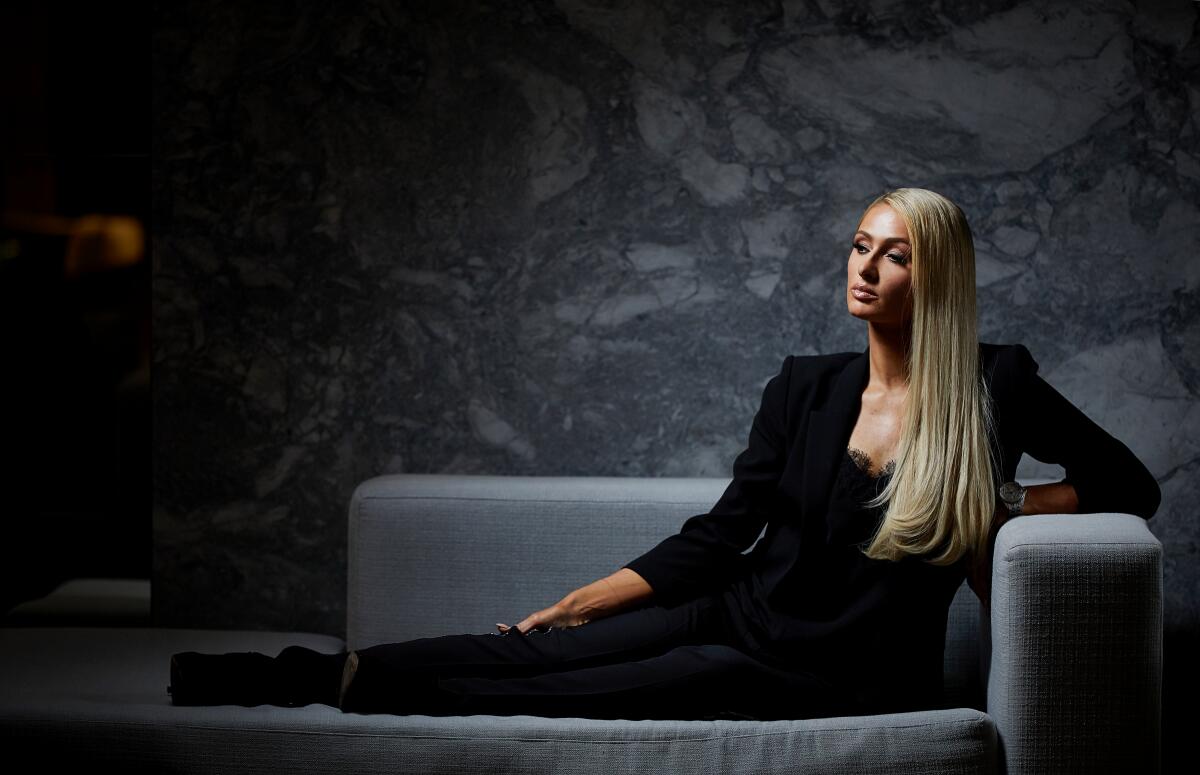  Paris Hilton wears a black suit while posing on a gray couch