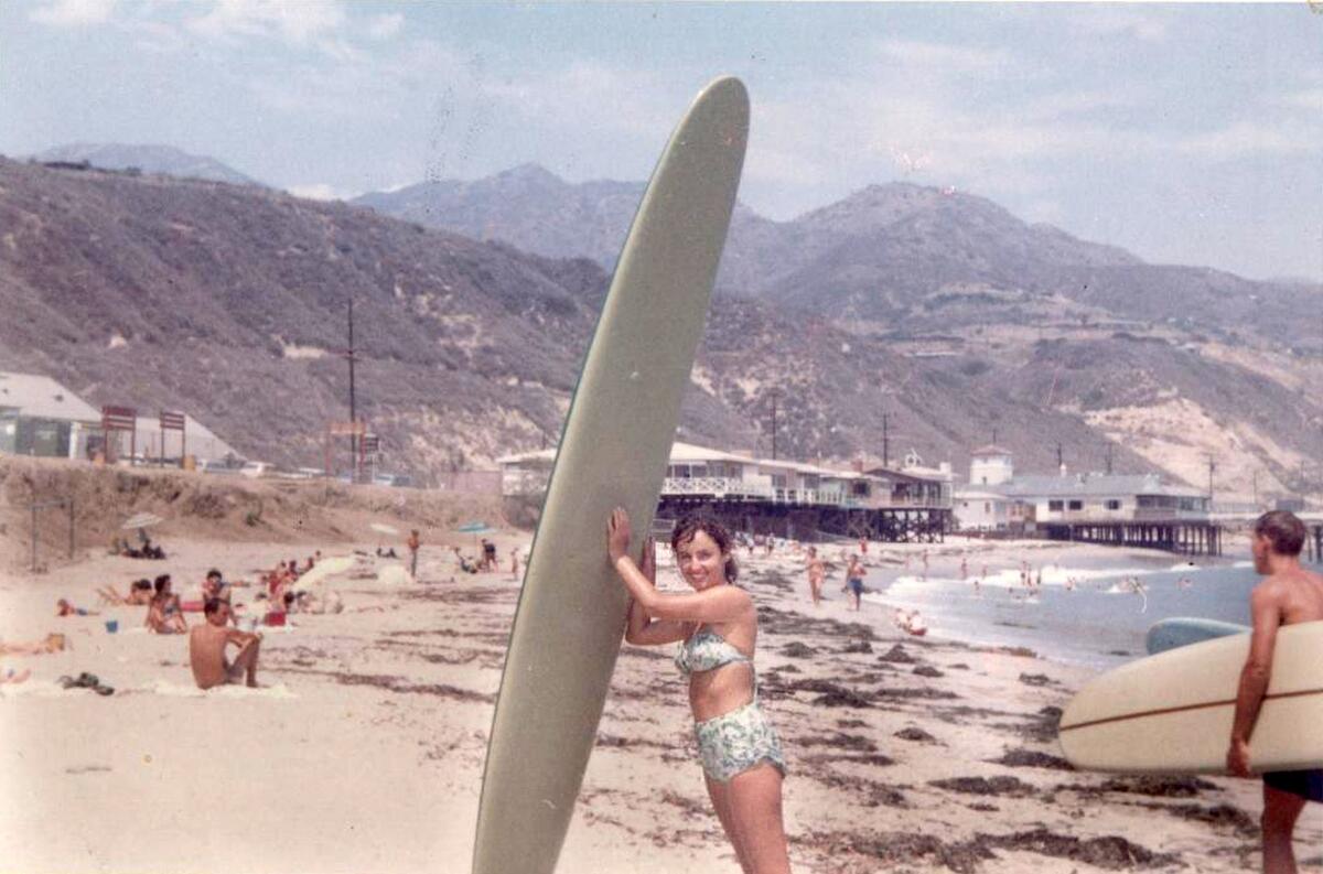 A woman with dark hair, in a bikini, holding up a large gray surfboard on a beach with hills as a backdrop