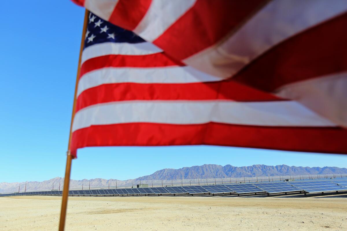 An American flag waves in a desert landscape with a large array of solar panels in the distance.