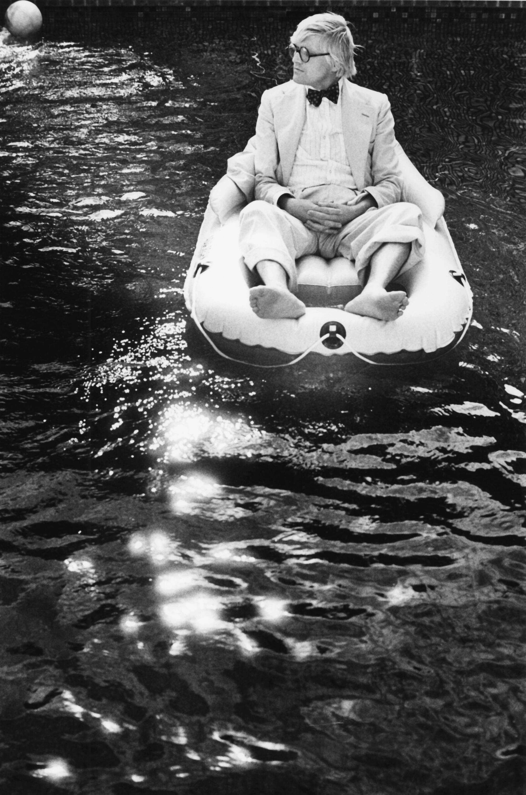 Artist David Hockney sits in an inflatable raft on a body of water.