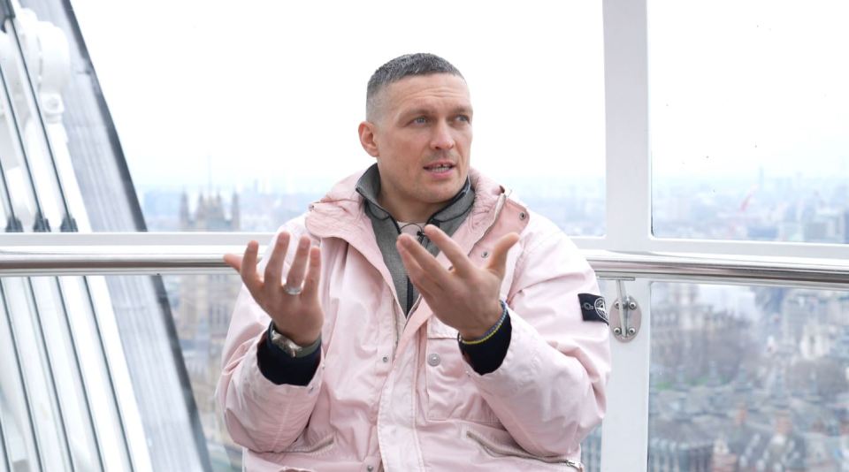 Usyk in a pink jacket being interviewed in a glass pod overlooking a city.