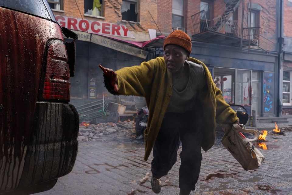 Film still of Lupita Nyong’o as Samira in A Quiet Place: Day One, running through a damaged city street toward a damaged vehicle.