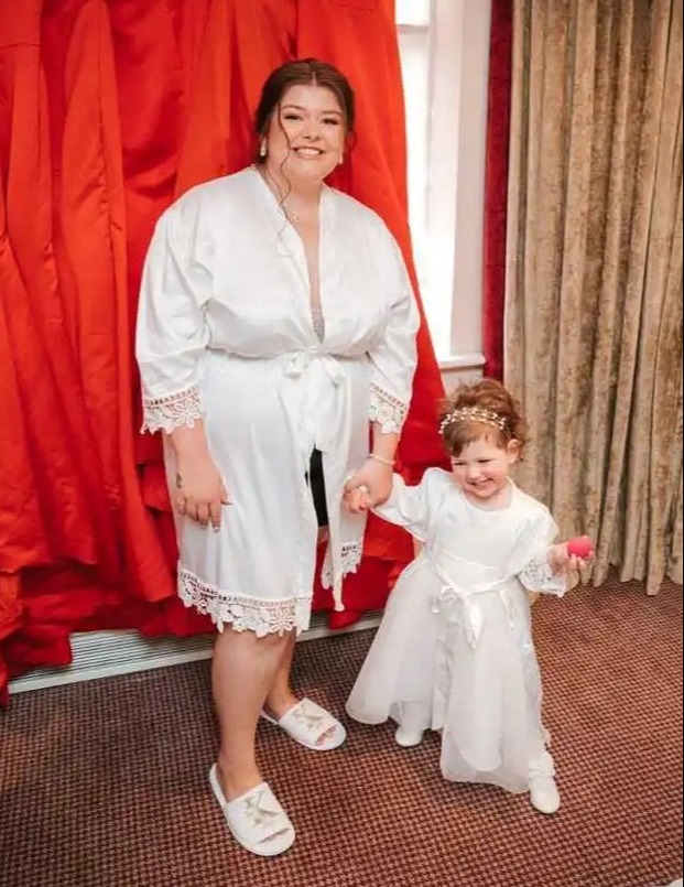 A bride and a young girl in white dresses stand in front of red bridesmaid dresses.