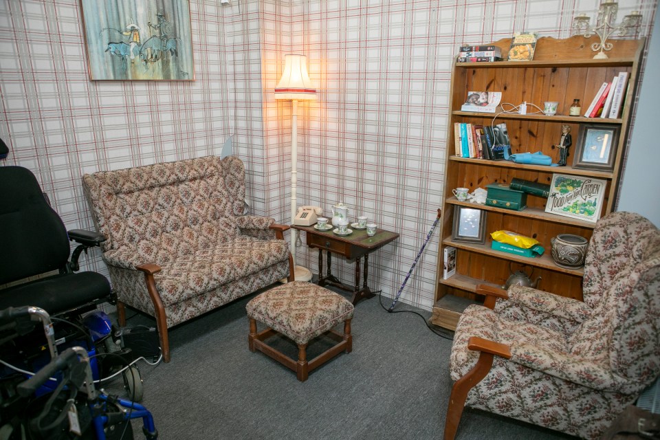 A living room with floral patterned furniture, a bookshelf, and a wheelchair.