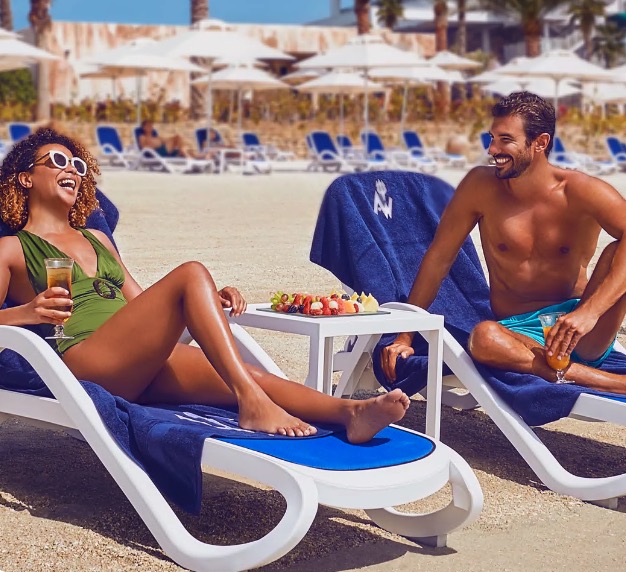 Couple relaxing on beach lounge chairs with drinks and snacks.