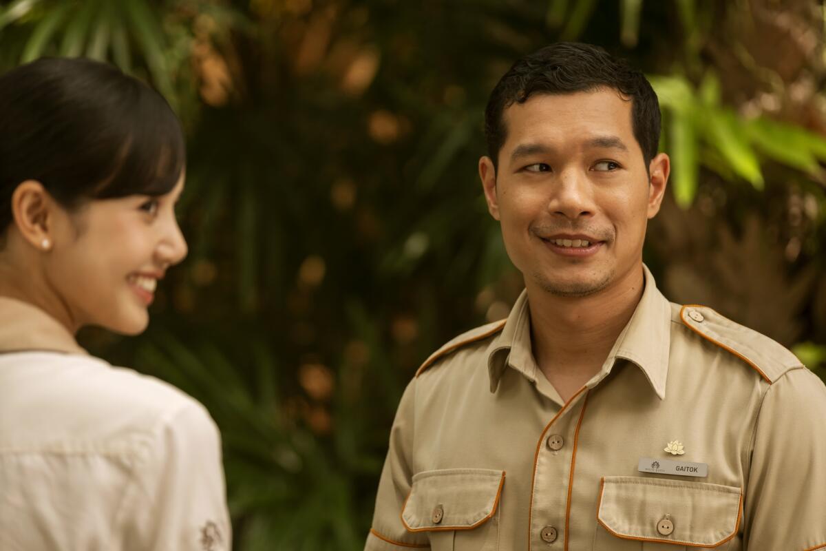 A smiling woman and a smiling man stand outdoors, both looking off in the same direction.