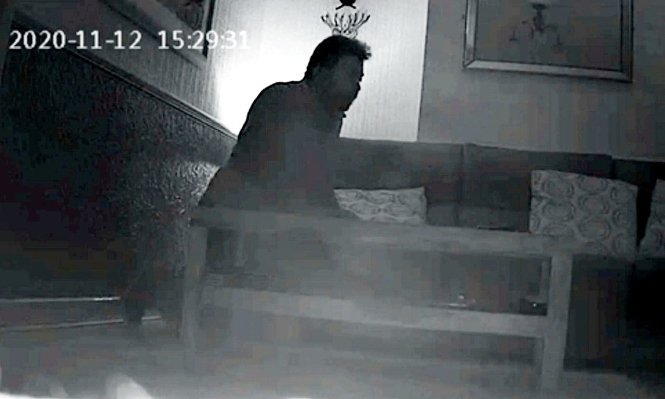 Low-light security camera image of a man placing a recording device under a coffee table.