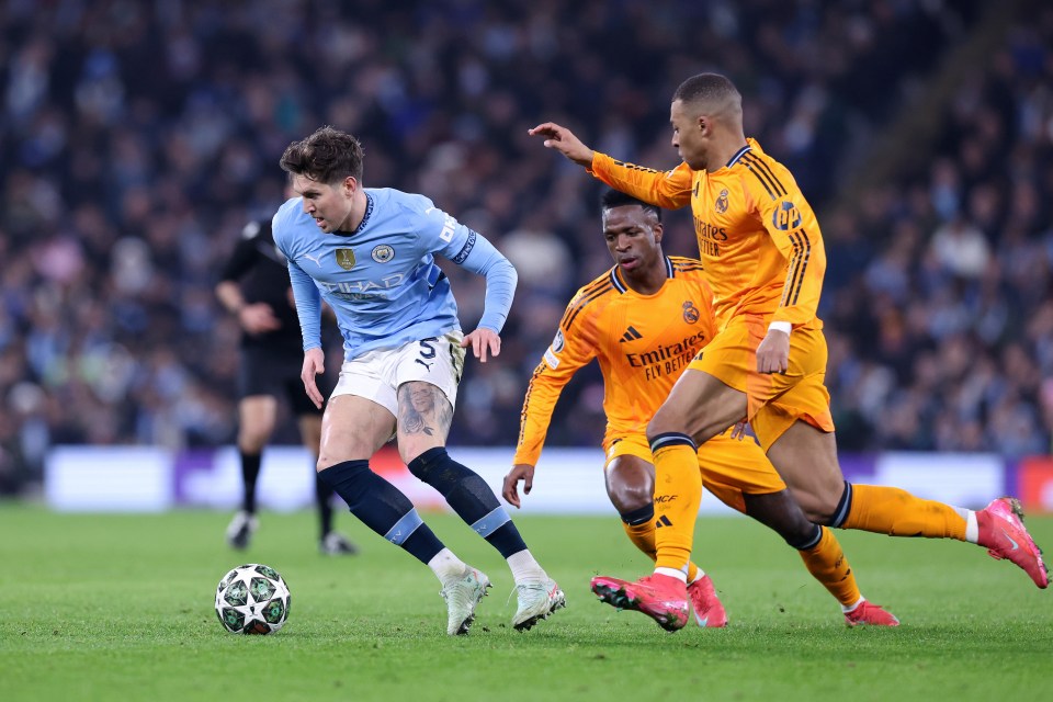 John Stones of Manchester City dribbling the ball past Vinicius Junior and Kylian Mbappé of Real Madrid.