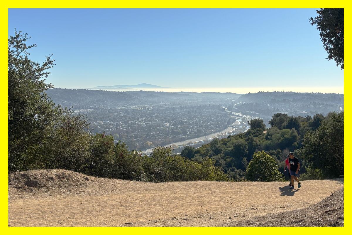 Two hikers on a dirt path pass an opening in the greenery. A highway stretches out in the distance.