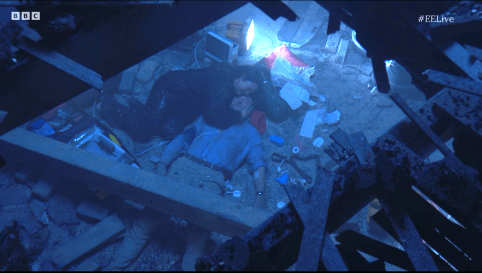 Screenshot of two people lying amidst rubble, illuminated by a lamp.