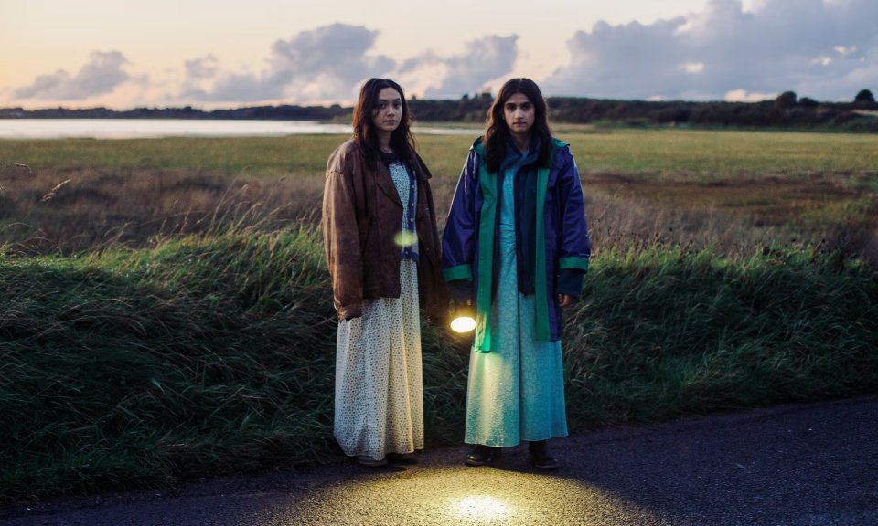 Film still of two girls standing on a road at dusk, holding a flashlight.