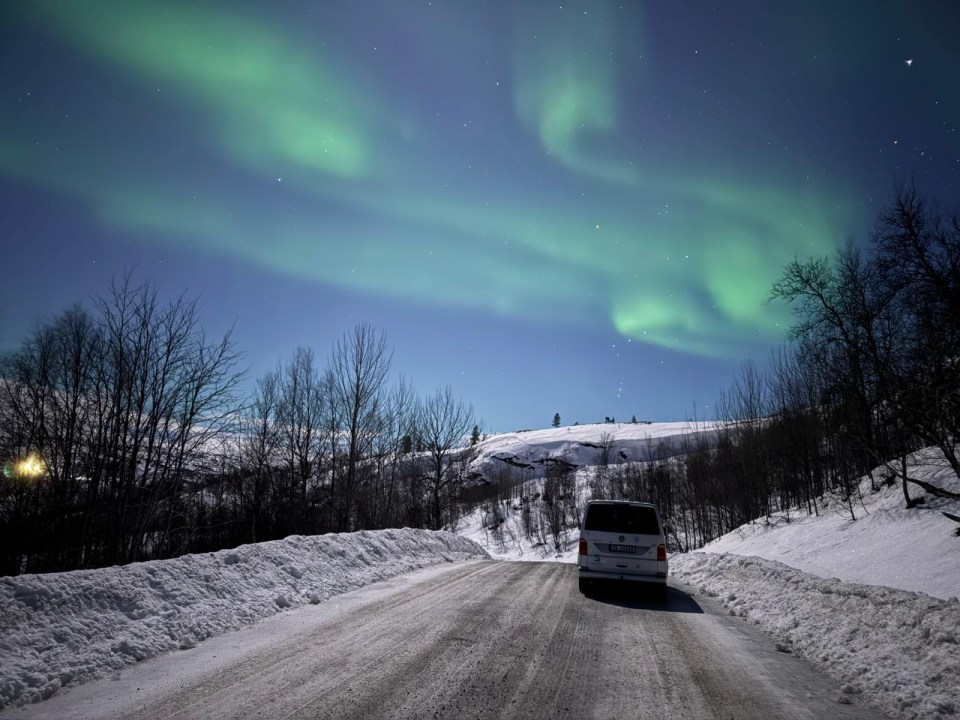 Van driving on a snow-covered road at night under the Northern Lights.