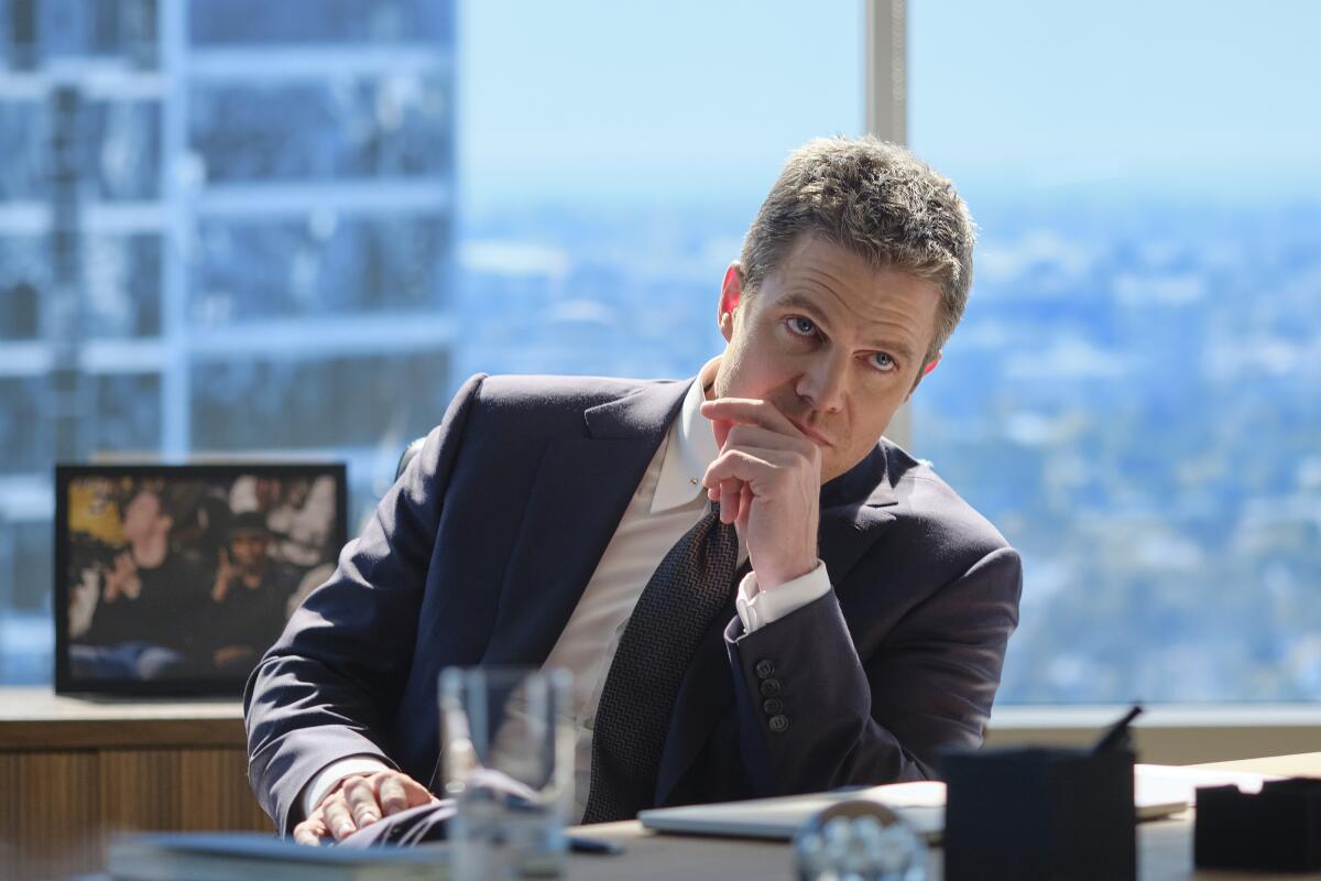 A man in a suit with his hand to his mouth sitting at a desk in high-rise building overlooking a cityscape.