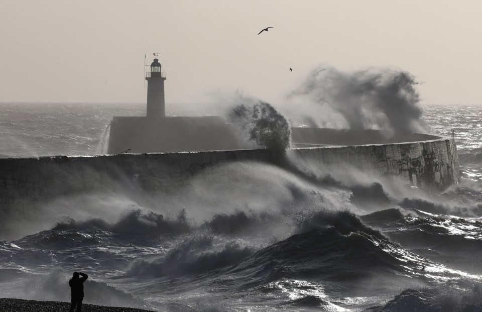 Person photographing large waves crashing against a breakwater with a lighthouse during a storm.