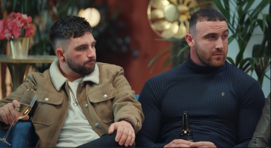 Two men sitting on a couch, one holding a glass of champagne, the other holding a beer bottle.