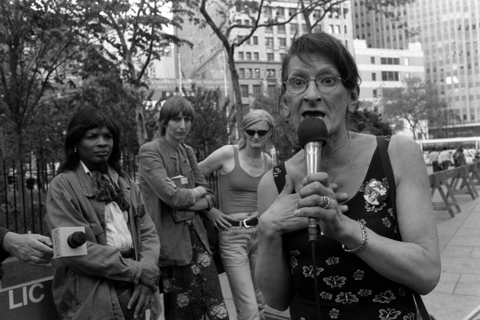 Black and white photo of Sylvia Rivera speaking at a rally, with Chelsea Goodwin visible in the background.