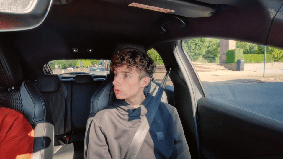 Screenshot of a young man looking surprised while sitting in the passenger seat of a car.