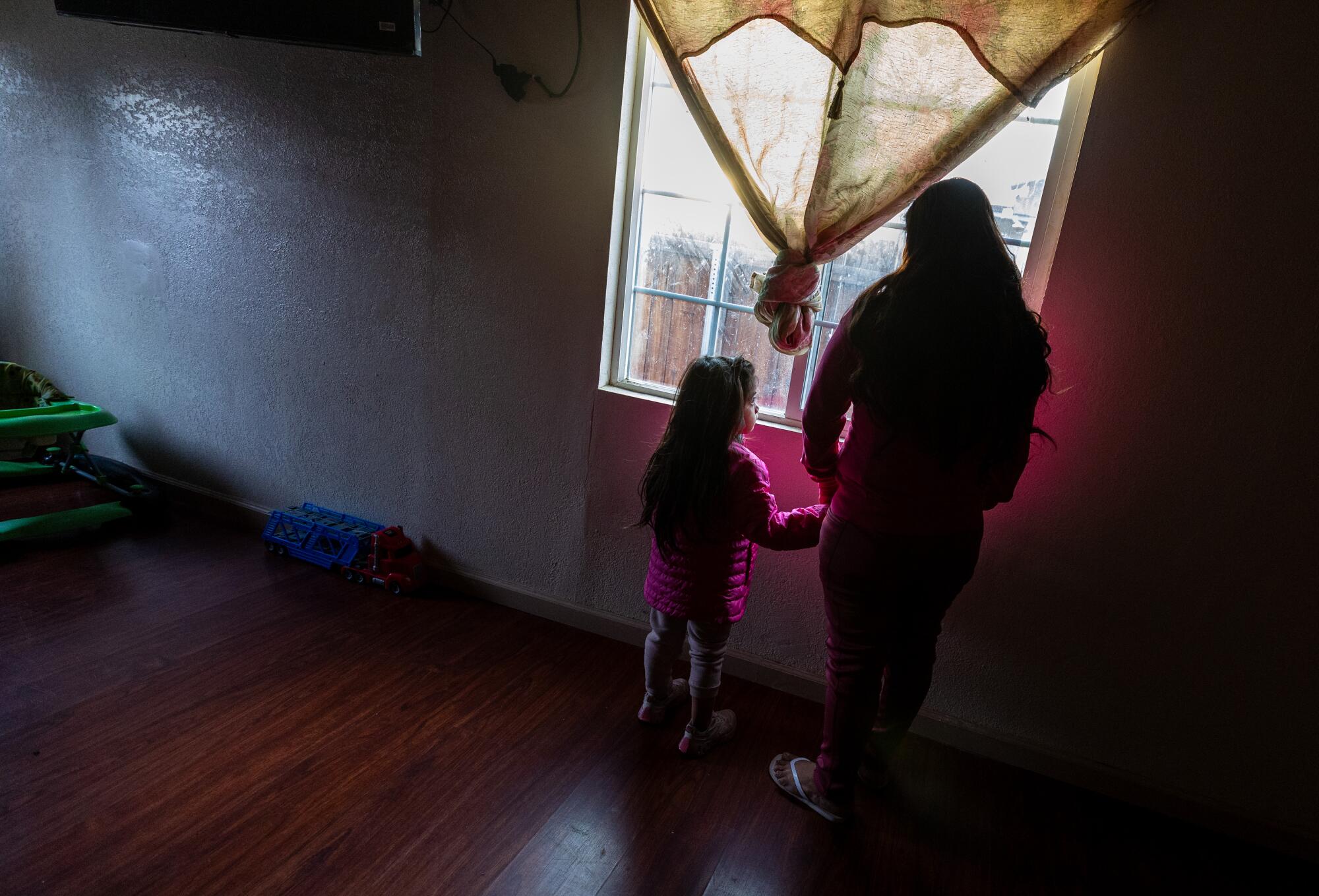 A woman and child look out a window in a sparsely furnished home. 