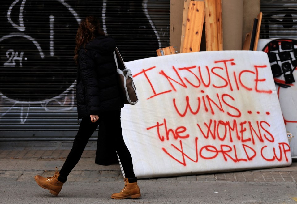 Woman walking past graffiti that reads "Injustice wins... the Womens World Cup" on mattresses.