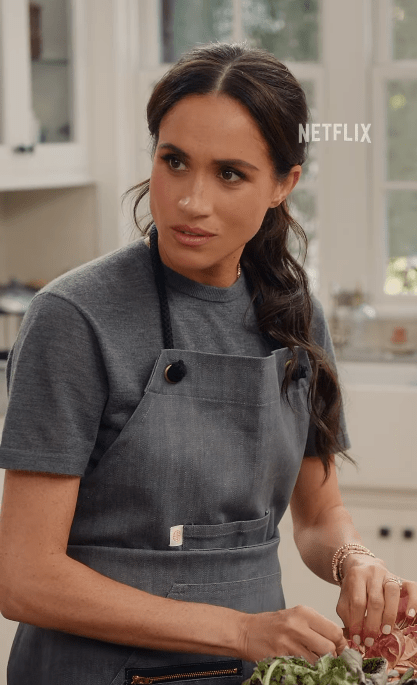 Meghan Markle wearing a gray apron, preparing food in a kitchen.