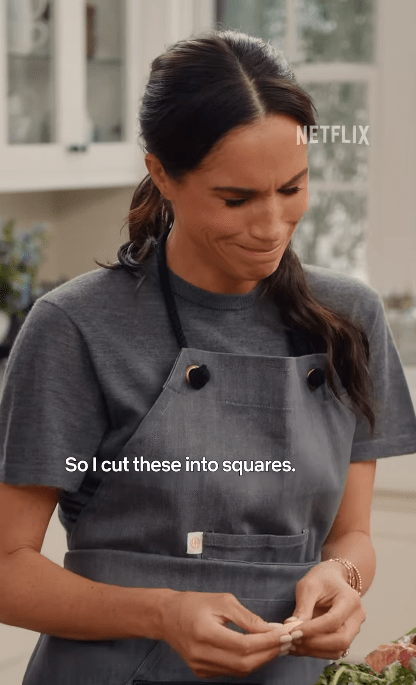 Screenshot of Meghan Markle in an apron, cutting something into squares.