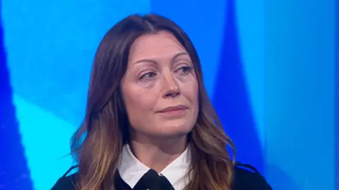 Mel Popham is in the Newsnight studio. She has long brown wavy hair and is wearing a dark coloured jacket with brass buttons and a white blouse.