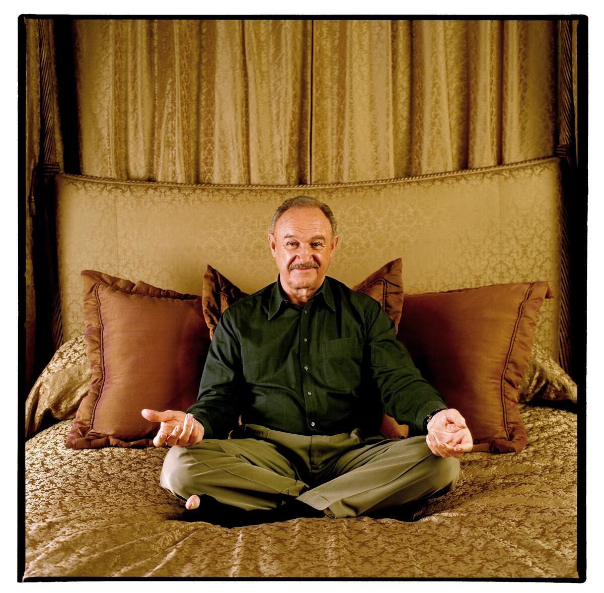 Gene Hackman sits cross-legged on a bed with his palms open facing up and resting on his knees.