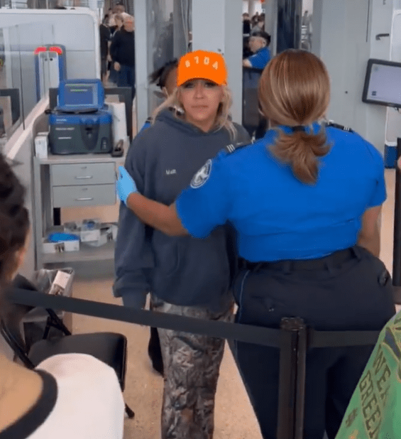 Woman in orange hat being escorted by TSA agent through airport security.