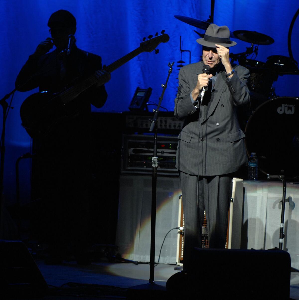 Leonard Cohen, onstage in a dark suit, holds a microphone in one hand and tips his hat with the other.