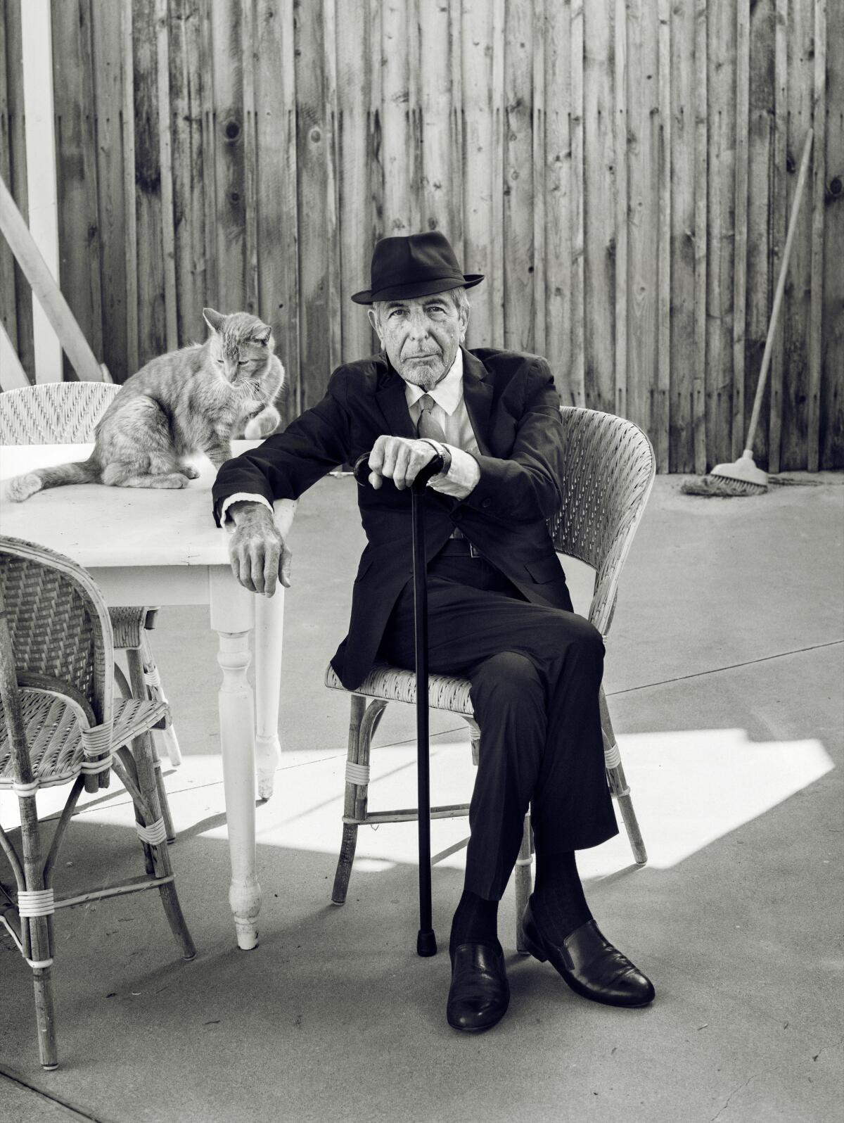 A man in dark suit and hat sits next to a table on which sits a cat