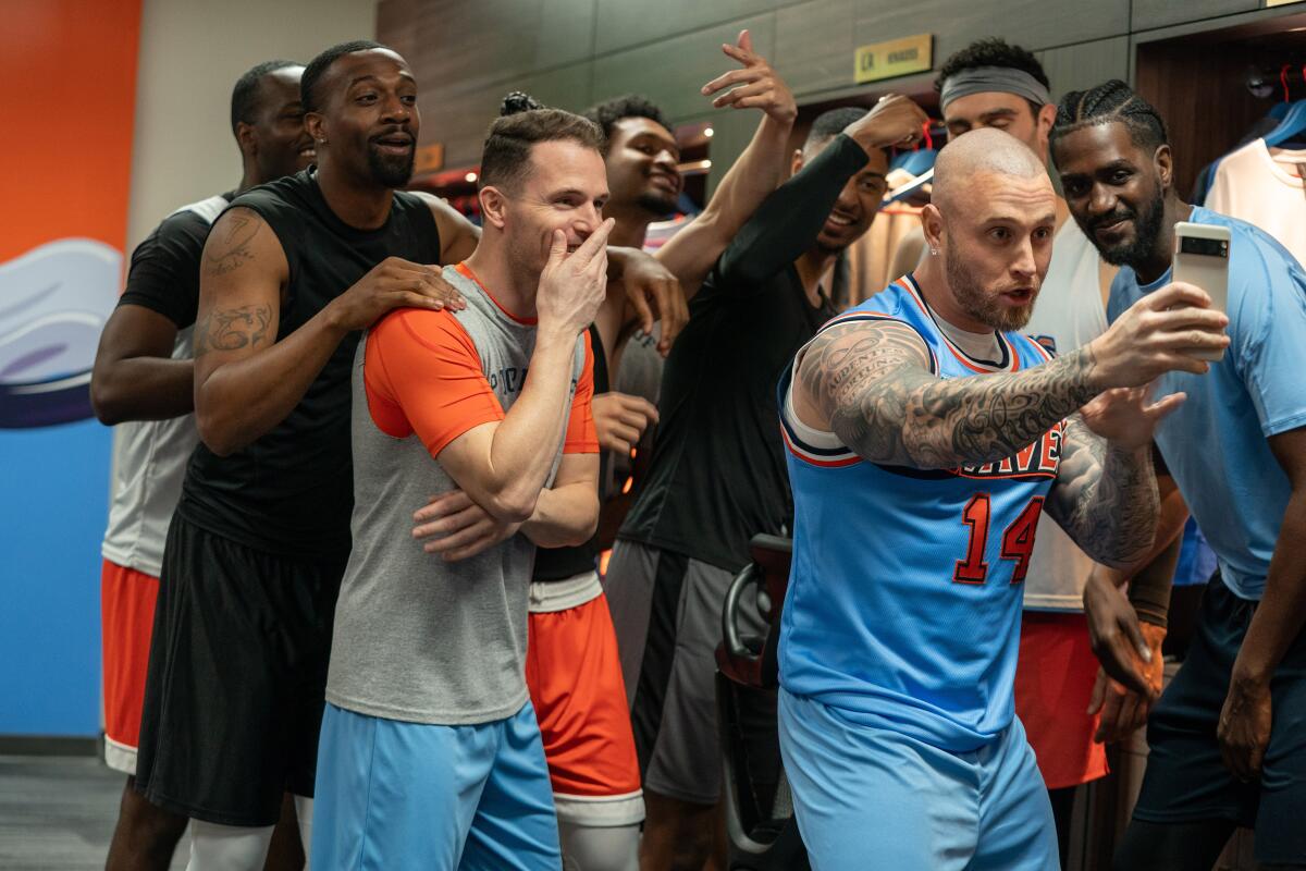 A group of men in T-shirts and basketball jerseys crowd around a man holding up a cellphone.