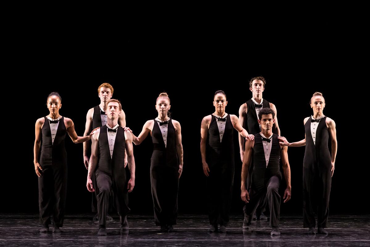 Eight men and women from Twyla Tharp Dance in a rough line onstage in sleek tuxedo-like costumes.