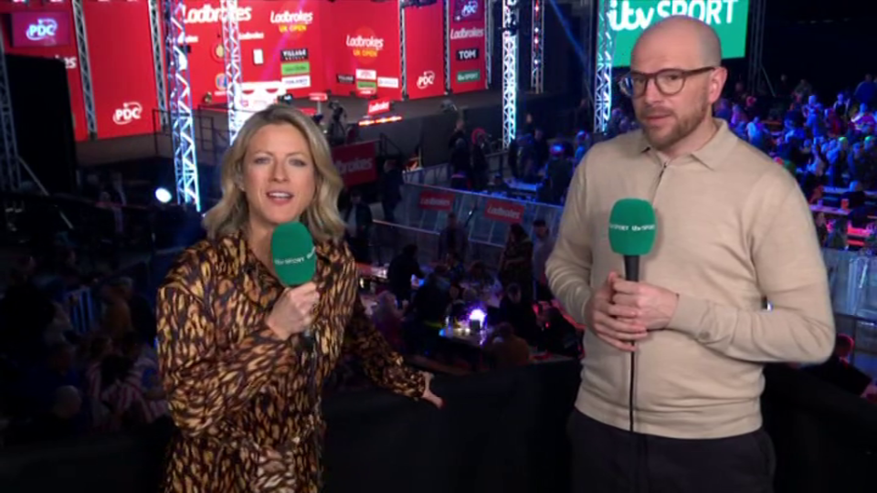 Two sports reporters holding microphones at a darts tournament.