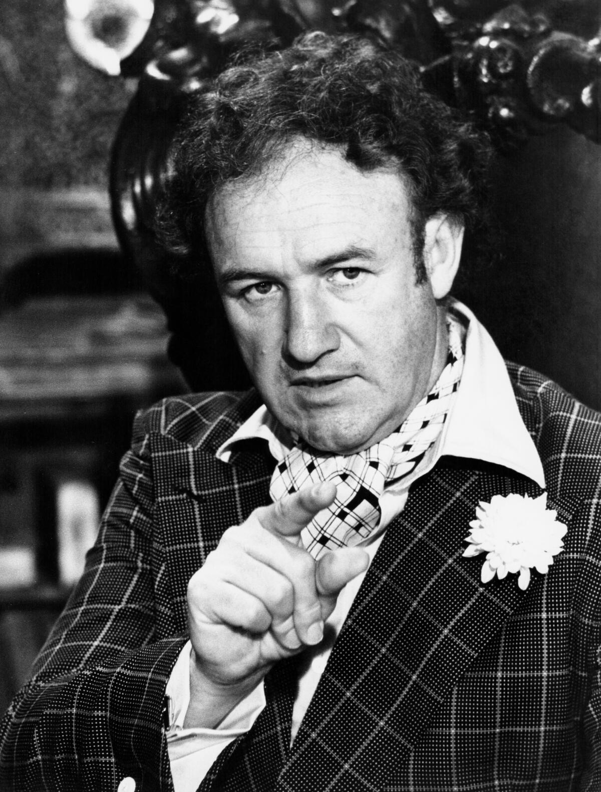 Gene Hackman points a finger. He has curly hair and wears a windowpane check suit with boutonniere and cravat.