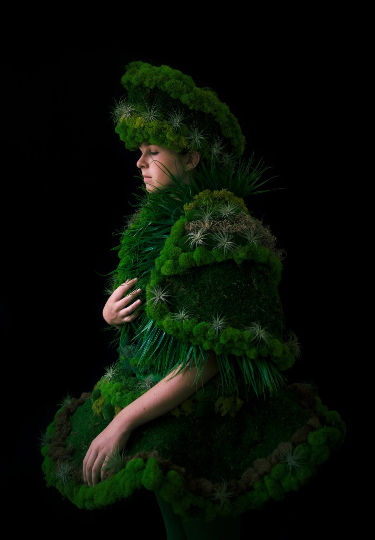 A woman clothed in a hat and coat made from various shades of moss. 