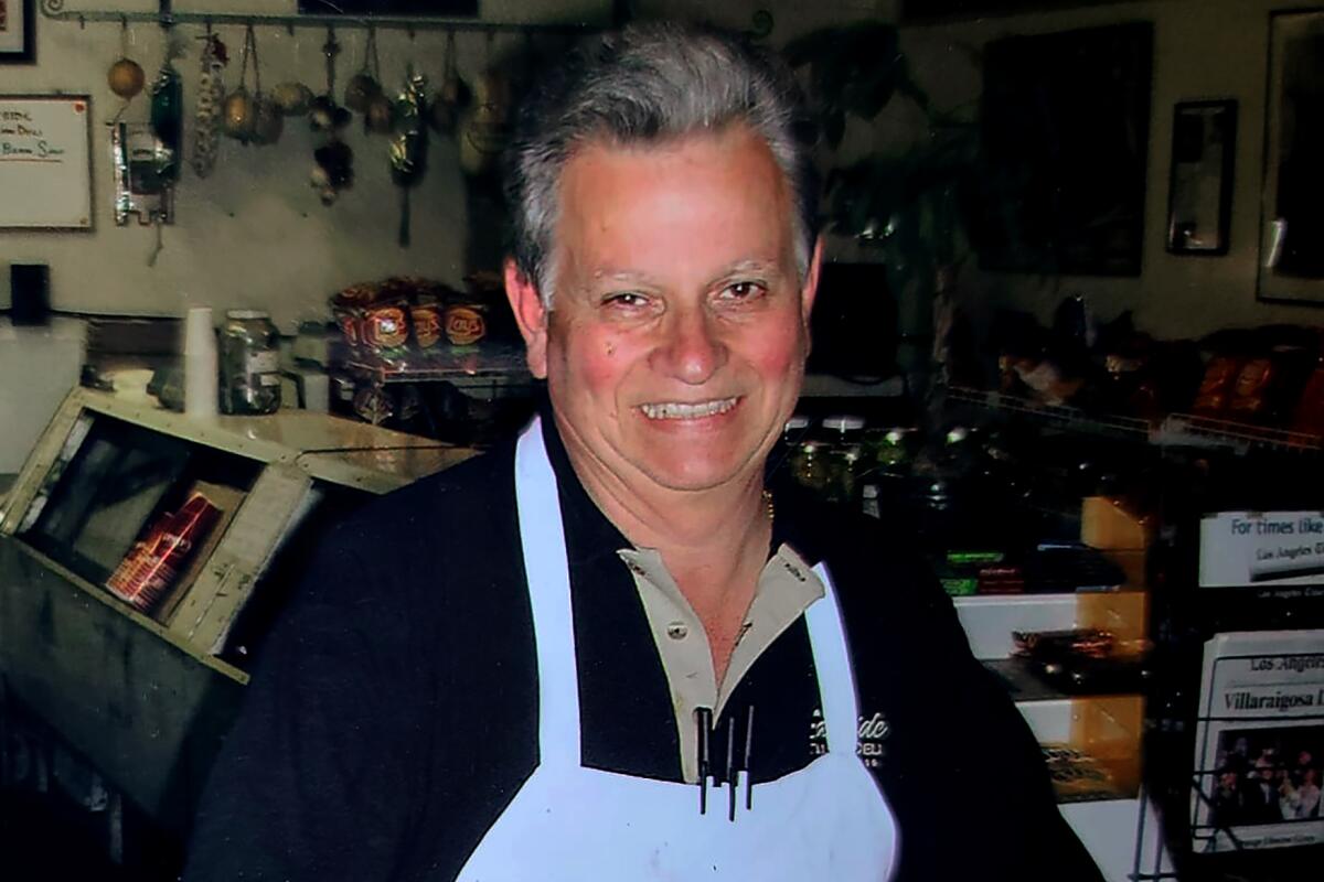 A smiling, gray-haired man in a blue apron stands behind a wooden counter. 