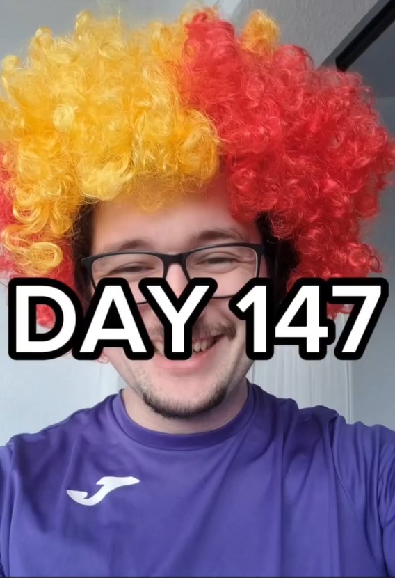 Man in a red and yellow wig, glasses, and purple shirt; text overlay: "DAY 147".