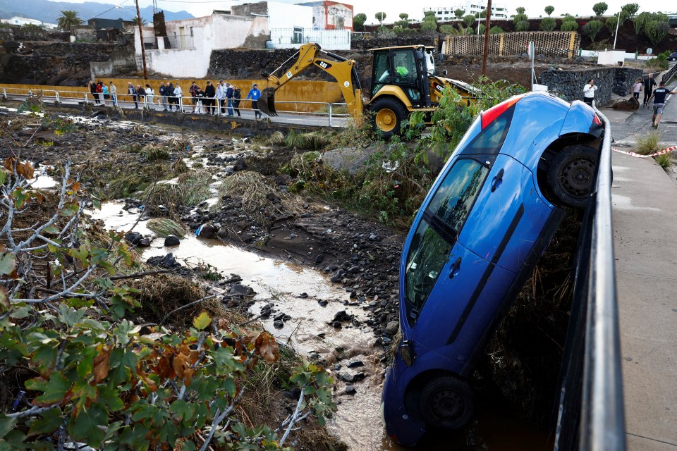 A blue car swept away by floodwaters rests against a railing; onlookers watch from a nearby bridge.