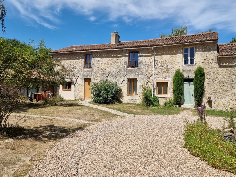 Stone house with multiple windows and doors, surrounded by a gravel driveway and landscaping.