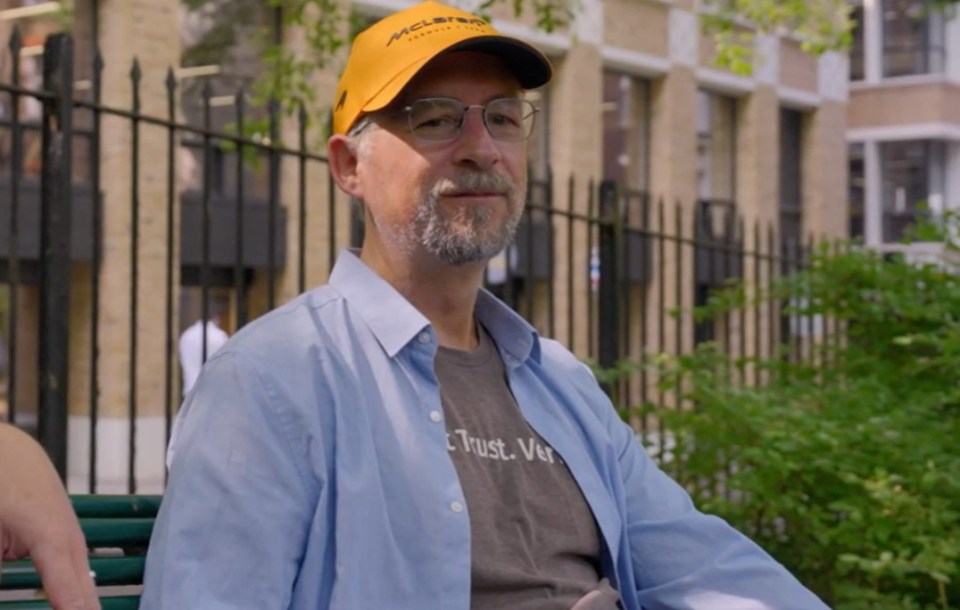 Man wearing a light blue shirt and orange baseball cap sits on a park bench.