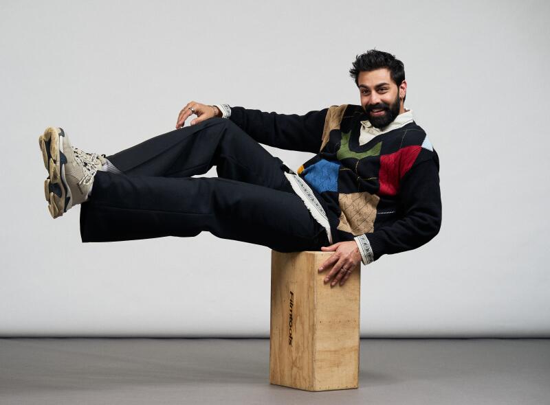 A man in a blue argyle sweater and slacks sits sideways on a wooden box.