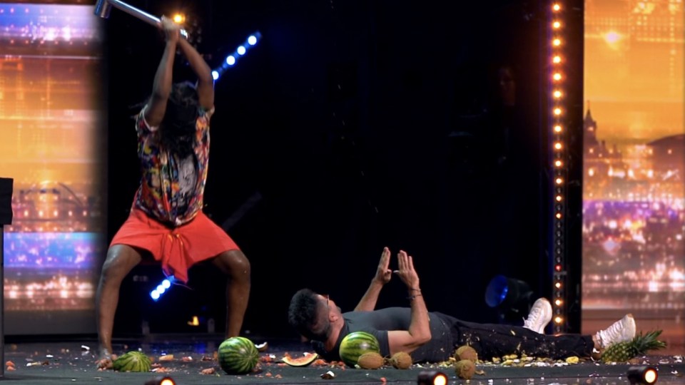 A person swinging an axe at watermelons and coconuts while another person lies on the ground.