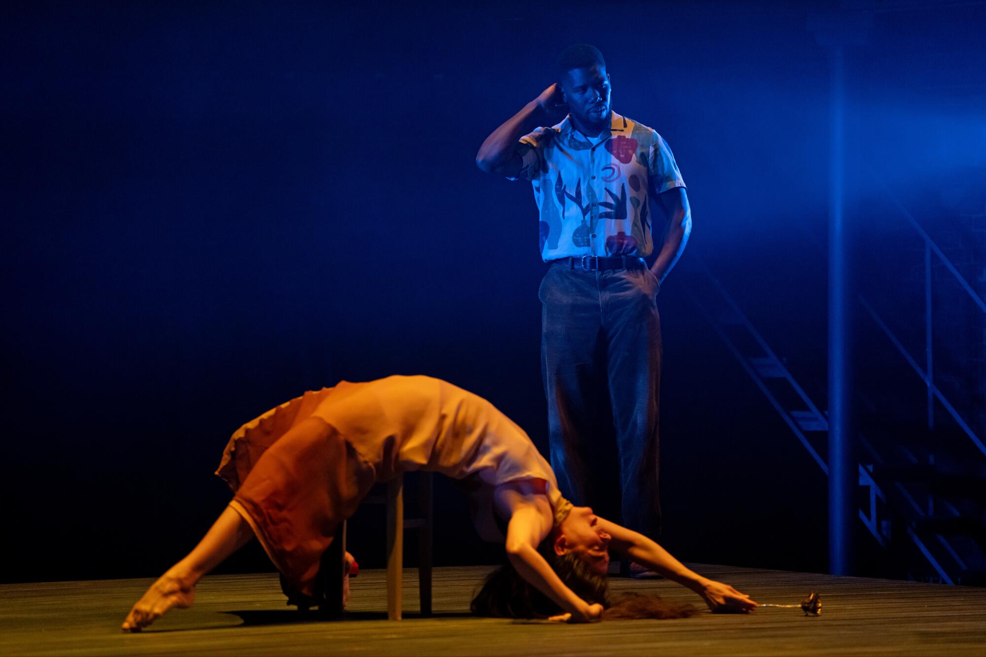 A man lighted in blue stands upstage of a woman lighted in orange who looks up as she arches gracefully over a table