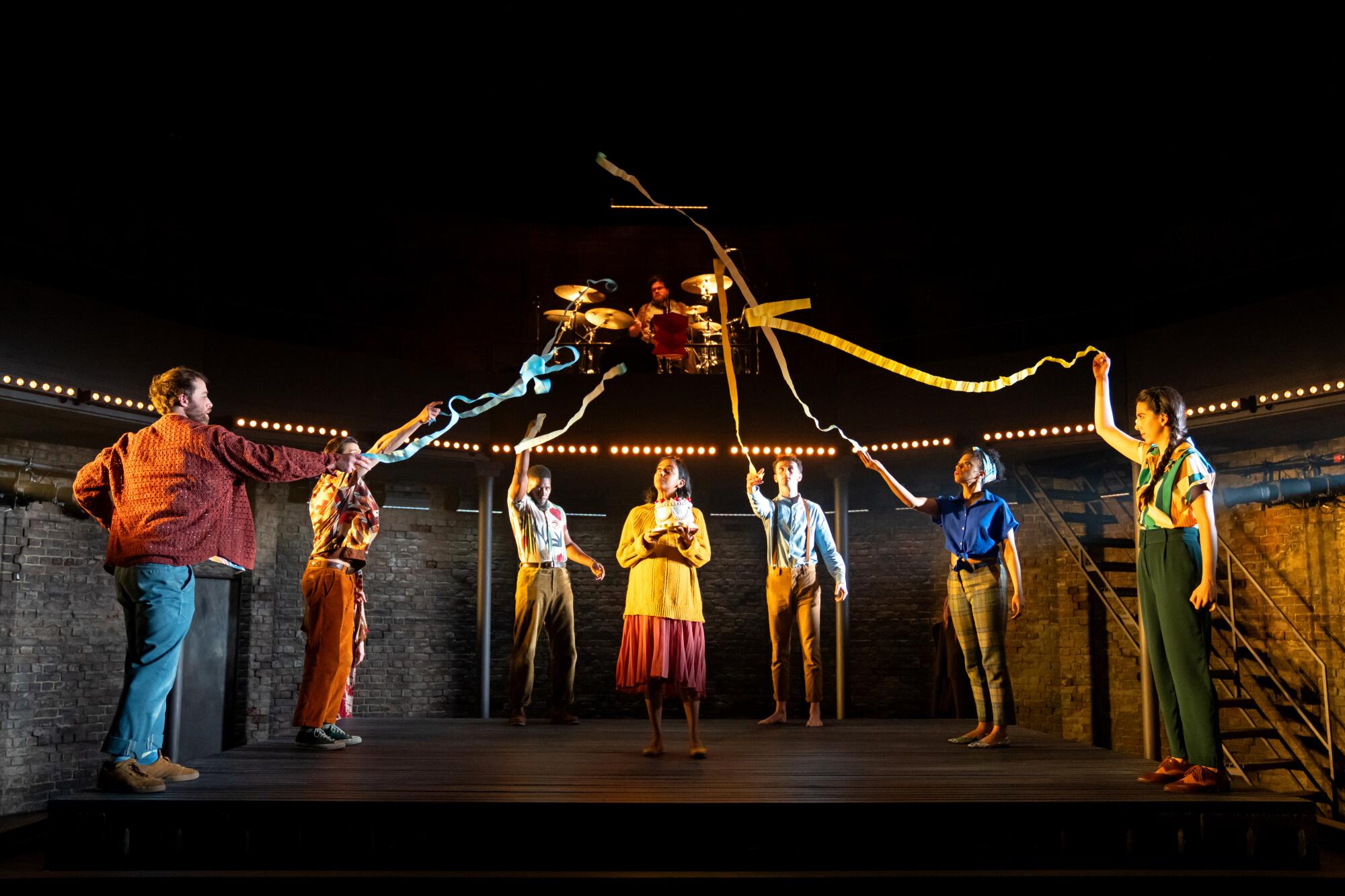 Seven actors stand onstage in a semicircle, holding up streamers attached to a ceiling fan